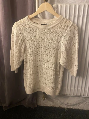 Vit stickad topp från Pieces - Säljer en söt vit stickad topp från Pieces med korta puffärmar och ett detaljerat hålmönster. Tröjan har rund halsringning och ribbade kanter. Perfekt att styla med jeans eller kjol för en trendig look.