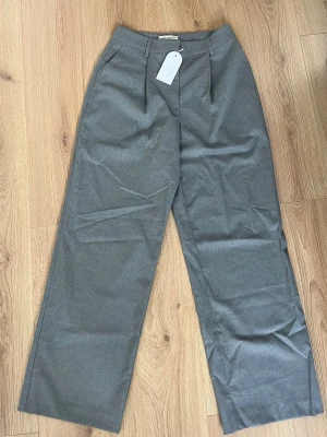 Grå kostymbyxa från Sanne Alexandra - Stilrena grå kostymbyxor från Sanne Alexandra med raka ben och pressveck. Byxorna har hällor för bälte och diskreta sidofickor. Perfekta för en clean och modern look. Materialet är slätt och ger en dressad känsla. Helt oanvända med lapp kvar