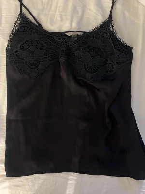 Svart spetslinne från H&M - Snyggt svart linne från H&M med tunna axelband och detaljerad spets upptill. Linnet har en mjuk, glansig finish och spetsen har blommönster som ger en elegant touch. Perfekt att styla lager på lager eller till jeans.