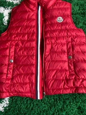 Röd väst från Moncler - Riktigt snygg röd väst från Moncler i princip oanvänd, väldigt bra skick. Box och kvitto finns! Storlek 12y, 152cm lång, passar upp till 14 år gammal. 