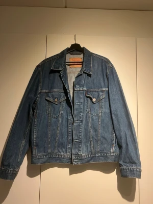 Vintage Levi's jeansjacka - Snygg blå jeansjacka från Levi's i klassisk modell med raka linjer, två bröstfickor med knappar och silverfärgade detaljer. Jackan har en tidlös design och är tillverkad i slitstarkt denimtyg. Perfekt för dig som gillar streetstyle och vintagevibbar.