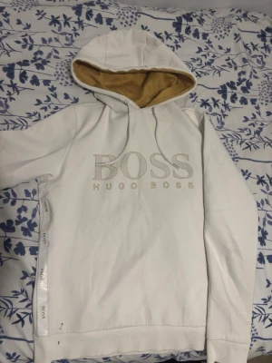 Vit hoodie från Hugo Boss - Snygg vit hoodie från Hugo Boss med stor logga framtill och beige insida på huvan. Klassisk passform med dragsko i huvan och ribbade muddar. Perfekt för en stilren och avslappnad look.              OBS!!! Som du ser på bilden nere till vänster så finns det små märken från en tuschpenna annars är den perfekt