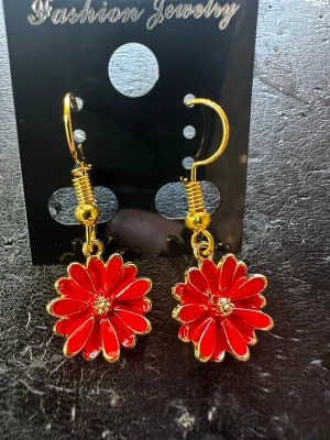 Röda blomörhängen med gulddetaljer - Snygga örhängen i form av röda blommor med guldfärgade kanter och detaljer. Hängande modell med krok, perfekt för att ge en färgklick till din stil. Små glittrande stenar i mitten av varje 