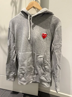 Grå Comme des Garçons PLAY hoodie - Säljer en grå hoodie från Comme des Garçons PLAY med det ikoniska röda hjärtat med ögon på bröstet. Hoodien har huva med dragsko, stor magficka och är gjord i mjukt bomullsmaterial. Perfekt för en chill och stilren look.