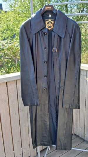 Svart lång läderrock orginal trenchcoat style. - Snygg svart lång läderrock trenchcoat style med klassisk krage, knäppning framtill och bälte i midjan. Rocken har raka ärmar och två fickor framtill. Denna rock är mycket gammal och stort samlar värde. Hoppas den kan komma till någon som uppskattar den.