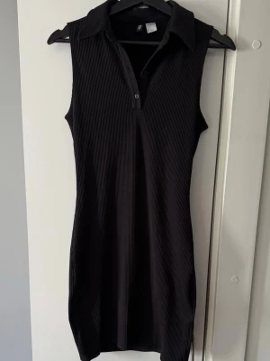 Svart ribbad klänning från H&M - Svart ribbad klänning från H&M med krage och knappar framtill. Ärmlös modell i kort längd, perfekt för en trendig och stilren look. Materialet är mjukt och stretchigt för en bekväm passform.