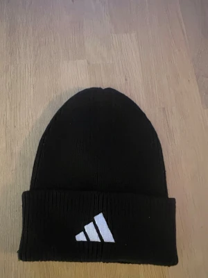 Svart ribbstickad mössa från Adidas - Svart ribbstickad mössa från Adidas med uppvikt kant och vit logga framtill. Klassisk och enkel design som passar perfekt till kalla dagar. Tillverkad i mjukt och stretchigt material för skön passform.