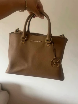 Beige handväska från Michael Kors - Väskan är i fint skick. Finns en liten reva (se bild) som säkert går att sy ihop, inget som påverkar användningen 