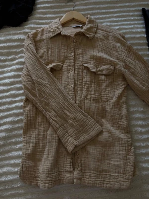 Beige skjorta från ASOS DESIGN - Luftig beige skjorta från ASOS DESIGN i mjukt bomullstyg. Skjortan har lång ärm, två bröstfickor med lock och knappar framtill. 