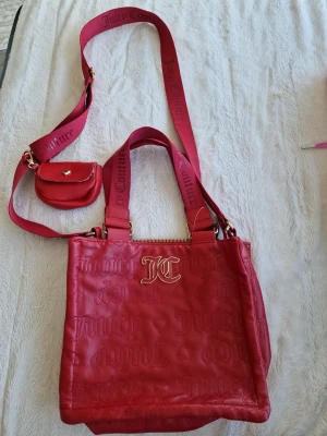 Röd Juicy Couture handväska - Snygg röd handväska från Juicy Couture i konstläder med JC-logga i guld framtill. Väskan har präglat mönster med Juicy Couture-text och hjärtan, bred axelrem med logga samt en liten matchande miniväska på remmen. Perfekt för dig som vill sticka ut!