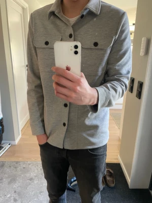 Ljusgrå overshirt med svarta knappar - Snygg ljusgrå overshirt från Only&Sons med klassisk krage och svarta knappar framtill. Två stora bröstfickor med lock och knapp. Rak passform och långa ärmar. Perfekt att slänga över en t-shirt för en clean look.