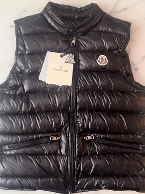 Svart dunväst från Moncler - Snygg svart dunväst från Moncler med glansig finish och klassisk logga på bröstet. Västen har två dragkedjeförsedda fickor framtill och hög krage. Perfekt för lager på lager och streetstyle. Materialet är dunfyllning med yttertyg i nylon.