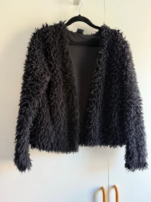 Svart fluffig teddyjacka - En svart teddyjacka med lång luddig pälsimitation och öppen front. Jackan har lång ärm och en rak, något kortare passform. Perfekt för dig som vill ha en mysig och trendig look. Materialet är mjukt och ger en cool statement-stil. Obs katt finns i hem!