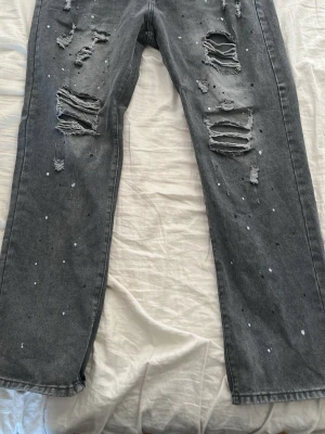 Grå slitna jeans med färgstänk - Snygga grå jeans med raka ben, stora slitningar och hål framtill samt vita färgstänk över hela byxan. 
