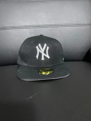 Svart New York Yankees keps - Snygg svart keps från New Era med klassiska New York Yankees-loggan broderad i vitt framtill. Kepsen har rak skärm och sticker ut med sin sportiga stil. Perfekt accessoar för dig som gillar streetwear och baseball.