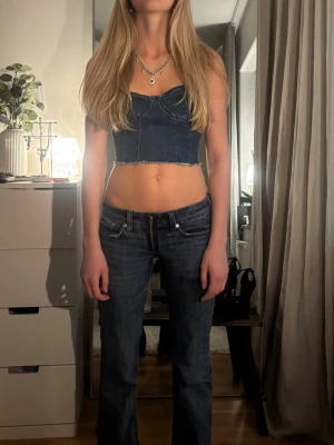 Mörkblå jeans denim korsett - Trendigt croppad korsettopp i mörkblå denim med rå kant nertill och markerade sömmar. Toppen har smickrande passform och tunna axelband. Perfekt att styla med jeans eller kjol för en cool look.