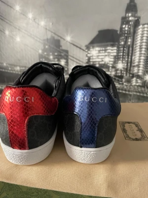 Gucci sneakers med röd och blå detalj - Snygga Gucci sneakers med svart canvas och GG-mönster, samt metallicröd detalj på ena hälen och metallicblå på den andra. Skorna har vita sulor och svarta läderdetaljer. Lyxig känsla och ikonisk design från Gucci.