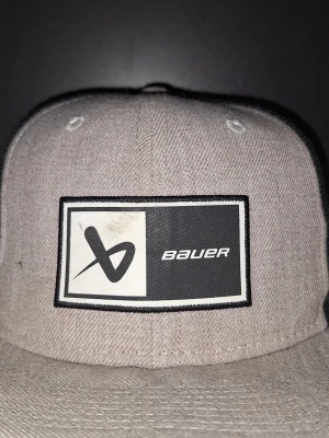 Grå och svart keps från Bauer - Snygg keps från Bauer i grått tyg framtill och svart mesh baktill. Kepsen har en rak skärm och en stor patch med Bauer-loggan på framsidan. Perfekt för en sportig och avslappnad stil.