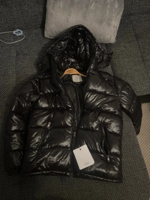 Svart glansig dunjacka från Moncler - Snygg svart dunjacka från Moncler med glansigt material och hög krage. Jackan har en klassisk puffer-design, dragkedja framtill och Moncler-logga på ärmen. Perfekt för kalla vinterdagar och riktigt trendig look.