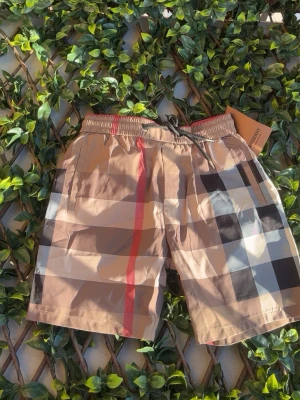 Beige rutiga shorts från Burberry - Snygga beige shorts från Burberry med klassiskt rutigt mönster i svart, vitt och rött. Shortsen har elastisk midja med snörning och är perfekta för sommaren. Ikonisk design som ger en trendig och lyxig känsla.