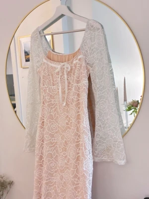 Vit spetsklänning med långa ärmar - Romantisk vit klänning med lång ärm i spets och vackert blommönster. Klänningen har en ljus nude underklänning och en söt knytning framtill vid halsen. Perfekt för dig som gillar en feminin och bohemisk stil.