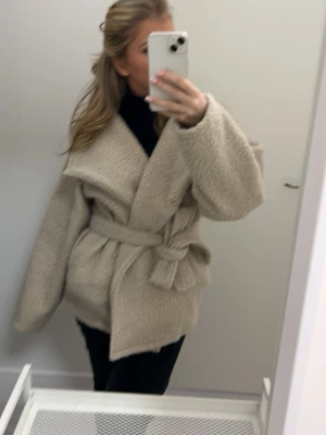 Beige oversized teddyjacka med skärp - Supermysig beige teddyjacka med oversized passform och bred krage. Jackan har ett matchande knytskärp i midjan som ger en snygg siluett. Perfekt för kalla dagar när du vill vara både varm och trendig. Materialet är fluffigt och mjukt för maximal komfort.