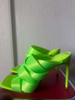 Neongröna pumps med flätade band - Säljer ett par trendiga neongröna pumps med höga stilettklackar och breda, flätade band över foten. Skorna har öppen tå och fyrkantig sula, vilket ger en modern vibe. Perfekta för dig som vill sticka ut med färg och stil.
