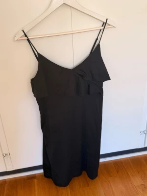 Svart kort klänning med volang H&M - Svart kort klänning med smala axelband och volangdetalj framtill från H&M. Klänningen har en enkel, stilren design och är perfekt för dig som gillar minimalistisk stil. Materialet känns lätt och svalt, vilket gör den skön att bära under varma dagar.