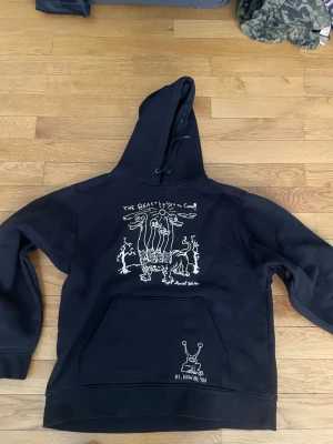 Svart hoodie med konstnärligt tryck - Cool svart hoodie med stor huva och unika vita konstmotiv av Daniel Johnston både fram och bak. Framtill finns en stor ficka och trycket visar fantasifulla figurer och texten 'The Beast'. Materialet ser ut att vara mjuk bomullsblandning. Perfekt för dig som gillar streetwear och konstnärliga plagg. Riktigt fet hoodie.