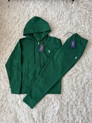 Grön tracksuit från Polo Ralph Lauren - Mysig grön tracksuit från Polo Ralph Lauren med huva och dragkedja på tröjan samt matchande byxor med resår i midjan. Båda delarna har den klassiska vita loggan broderad. Perfekt för chill dagar hemma eller när du vill vara bekväm och snygg.