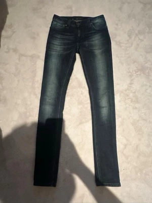 Mörkblå jeans från Nudie - Snygga mörkblå skinny jeans från Nudie med klassisk femficksdesign och diskreta slitningar framtill. Jeansen har en smal passform och är tillverkade i stretchigt denim för extra komfort. Perfekta för en trendig och avslappnad stil.