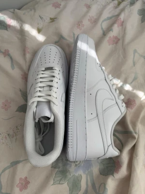 Nike Air Force 1  - Klassiska Nike Air Force 1 sneakers i helvitt läder med perforerad tå, vit sula och ikonisk swoosh på sidorna. Skorna har lågt skaft och vita skosnören. Perfekta för en clean och stilren look.