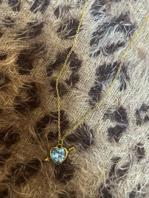 Halsband med hjärthänge och pil blå guld - Guldfärgat halsband med ett hjärformat hänge i blått glas och en liten pil genom hjärtat. Kedjan är tunn och elegant, och hänget har en glittrande effekt som fångar ljuset. Perfekt accessoar för att lyfta din outfit med en söt detalj. Rostfri