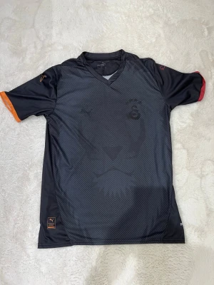 Svart fotbollströja från Puma - Snygg svart fotbollströja från Puma med diskret mönster och klubbmärke på bröstet. Tröjan har V-ringning, korta ärmar med orange och röd detalj på ärmsluten. Tillverkad i lätt och ventilerande funktionsmaterial, perfekt för träning eller match.