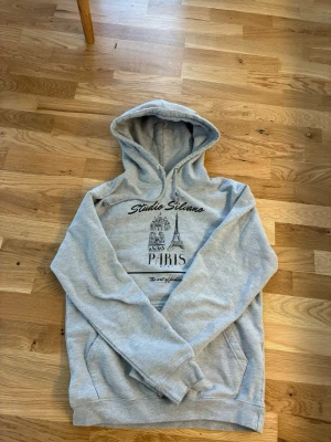Grå hoodie Studio Silano Paris S - Grå hoodie från Studio Silano med tryck av Eiffeltornet och texten Paris på bröstet. Klassisk modell med huva och känguruficka framtill. Mjuk bomullsblandning som är skön att ha på sig. Perfekt för en chill och avslappnad stil.