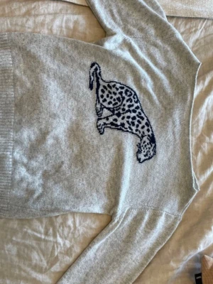 Ljusgrå stickad tröja med leopardmotiv SÖKES!!!!  - Ljusgrå stickad tröja med ett stort svart och vitt leopardmotiv på framsidan. Tröjan har långa ärmar och ribbstickade muddar vid ärmslut och nederkant. Perfekt för dig som gillar djurmönster och vill ha en unik detalj i garderoben.