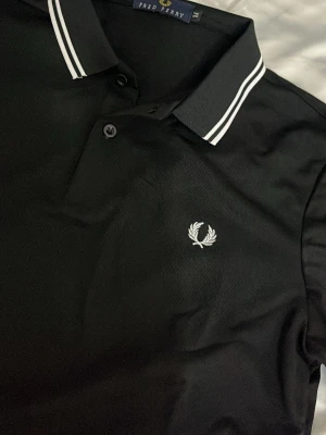 Svart pikétröja från Fred Perry - Klassisk svart pikétröja från Fred Perry med vit broderad logga på bröstet och vita ränder på krage och ärmslut. Tröjan har korta ärmar, knappslå och är gjord i ett mjukt bomullsmaterial. Perfekt för en clean och sportig look.