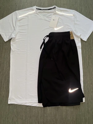 Nike träningsset t-shirt och shorts - Snyggt träningsset från Nike med en vit t-shirt och svarta shorts. T-shirten har rund hals och korta ärmar, medan shortsen har dragsko i midjan och reflekterande Nike-logga. Perfekt för gymmet eller löprundan. ⚠️ STORLEKSINFO: Lappen säger L (Asien), men passformen motsvarar EU Small (S). • Bröstvidd: 95–101 cm. • Midja: 88–94 cm. Snabba ryck! Först till kvarn! 🚀 