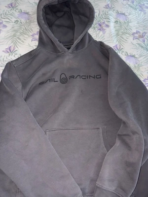 Grå hoodie från Sail Racing - Snygg grå hoodie från Sail Racing med svart logga på bröstet. Klassisk huva och stor magficka framtill. Perfekt för en chill och avslappnad stil. Mjuk insida och skön passform. Pris kan diskuteras vid snabb affär.