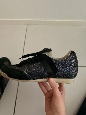 Svarta glitter sneakers från Serafini - Glittriga svarta sneakers från Serafini med svart mockadetalj vid tån och svart läderpanel på sidan. Skorna har svarta skosnören och en vit sula. Perfekta för dig som vill sticka ut med en unik och trendig look.