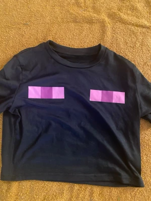 Svart croppad t-shirt med lila tryck - Cool svart croppad t-shirt med två lila rektangulära tryck framtill. Perfekt för dig som gillar en edgy och unik stil. Rund halsringning och korta ärmar. Passar till jeans eller kjol för en avslappnad look.