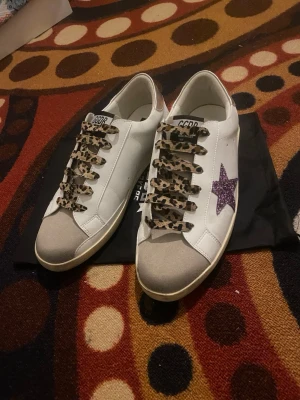 GGDB sneakers med leopardmönstrade snören - Snygga vita sneakers från Golden Goose med lila glittrig stjärna på sidan och leopardmönstrade skosnören. Skorna har grå tå och klassisk låg modell. Perfekta för dig som vill sticka ut med detaljer och cool design.