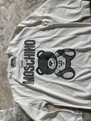 Vit Moschino sweatshirt med björn - Cool vit sweatshirt från Moschino med stort svartvitt tryck av en björn och texten 'This is not a Moschino toy'. Tröjan har rund hals och ribbade muddar. Perfekt statement-plagg för dig som gillar streetstyle och designer.