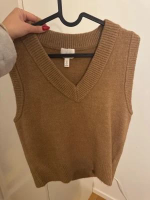 Beige stickad väst från H&M - Snygg beige stickad väst från H&M med v-ringad hals och breda ribbade kanter. Perfekt att styla över en skjorta eller t-shirt för en trendig lager-på-lager-look. Använd en gång