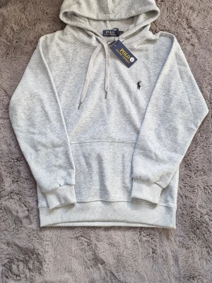Grå hoodie från Polo Ralph Lauren - Snygg ljusgrå hoodie från Polo Ralph Lauren med klassisk huva och dragsnören. Har en broderad logga på bröstet och en stor magficka framtill. Tillverkad i mjukt material som är skönt mot huden. Perfekt för en avslappnad och stilren look. 550kr först till kvarn