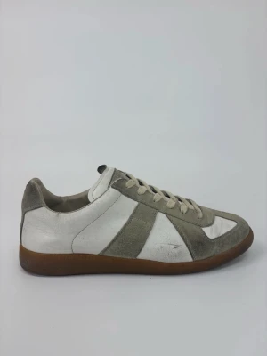 Vita och grå Maison Margiela sneakers med mockadetaljer - Snygga sneakers i vitt och grått med klassisk design och bruna gummisulor. Skorna har ovandel i läder och mocka, samt vita snören. Perfekta för dig som gillar stilrena och tidlösa sneakers med en retrovibe.