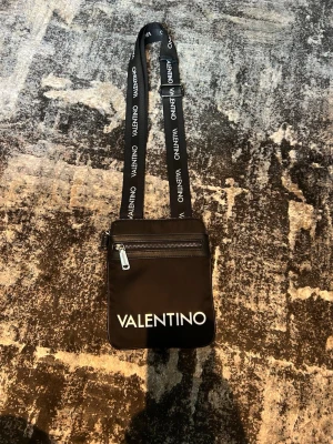 Svart axelväska från Valentino - Snygg svart axelväska från Valentino med vitt logotryck framtill och på axelremmen. Väskan har en dragkedja framtill och ett huvudfack med innerficka. Tillverkad i slitstarkt tygmaterial och har en justerbar axelrem.