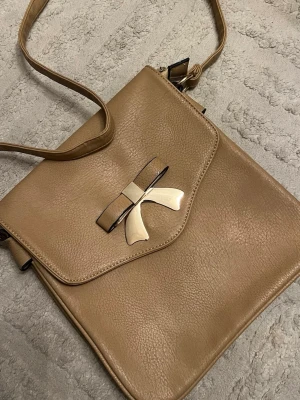 Beige axelväska med rosettdetalj - Snygg beige axelväska i konstläder med justerbar rem och lock med dekorativ metallrosett framtill. Väskan har en fyrkantig form och stängs med lock. Perfekt för dig som gillar stilrena accessoarer med en söt twist.