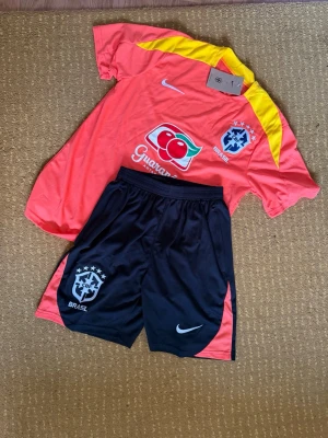Brasilien fotbolls träningsset - Säljer ett par svarta sportiga shorts och T shirt från Nike med Brasilien-logga och vita detaljer. Shortsen har orangea inslag på sidorna och är perfekta för fotboll eller träning. De har en avslappnad passform och är tillverkade i ett lätt och bekvämt material.
