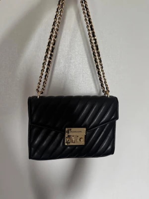 Svart axelväska Michael Kors - Snygg svart axelväska från Michael Kors i quiltat läder med guldfärgad kedjerem och vridlås framtill. Klassisk rektangulär form och elegant design med diskreta diagonala sömmar. Perfekt accessoar för att lyfta din outfit. När man öppnar låset är det lite skrapor som syns på bilden annars är den i fint skick. 